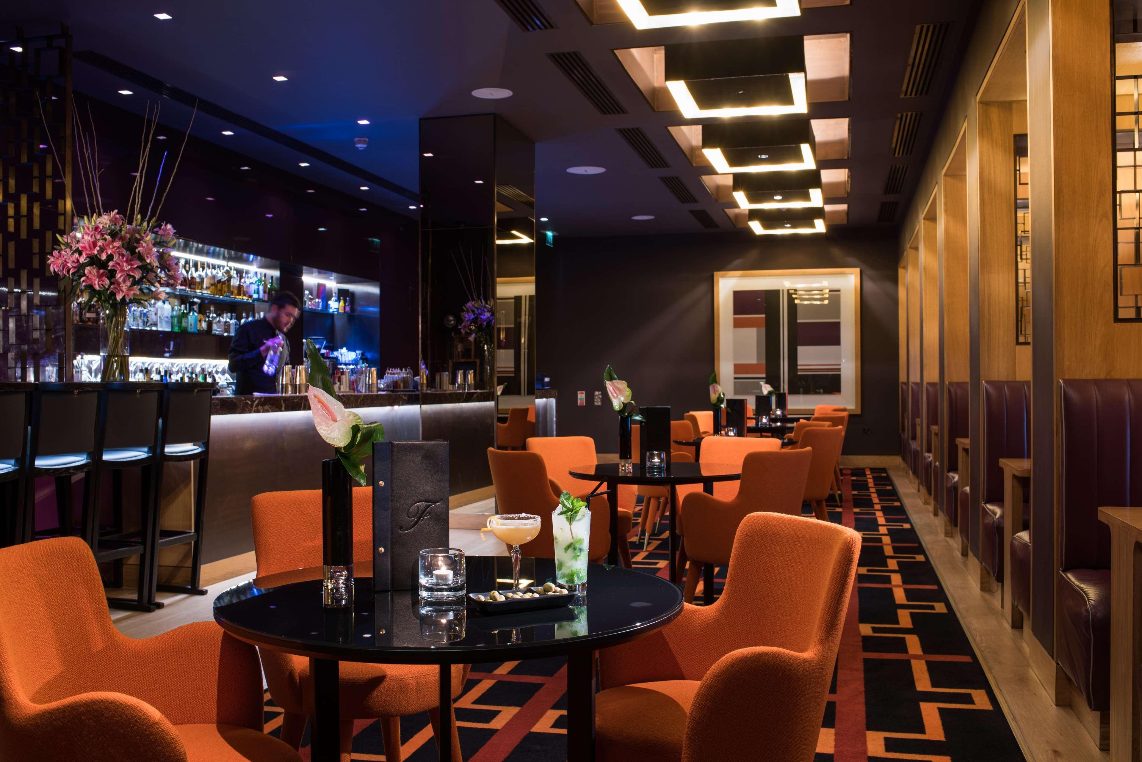 The Fitzwilliam Hotel Belfast Dış mekan fotoğraf The photo depicts a stylish bar or lounge area with a modern and sophisticated atmosphere. The space features dark walls accented by blue lighting and a sleek bar counter in the background, stocked with various drinks. The seating consists of round t