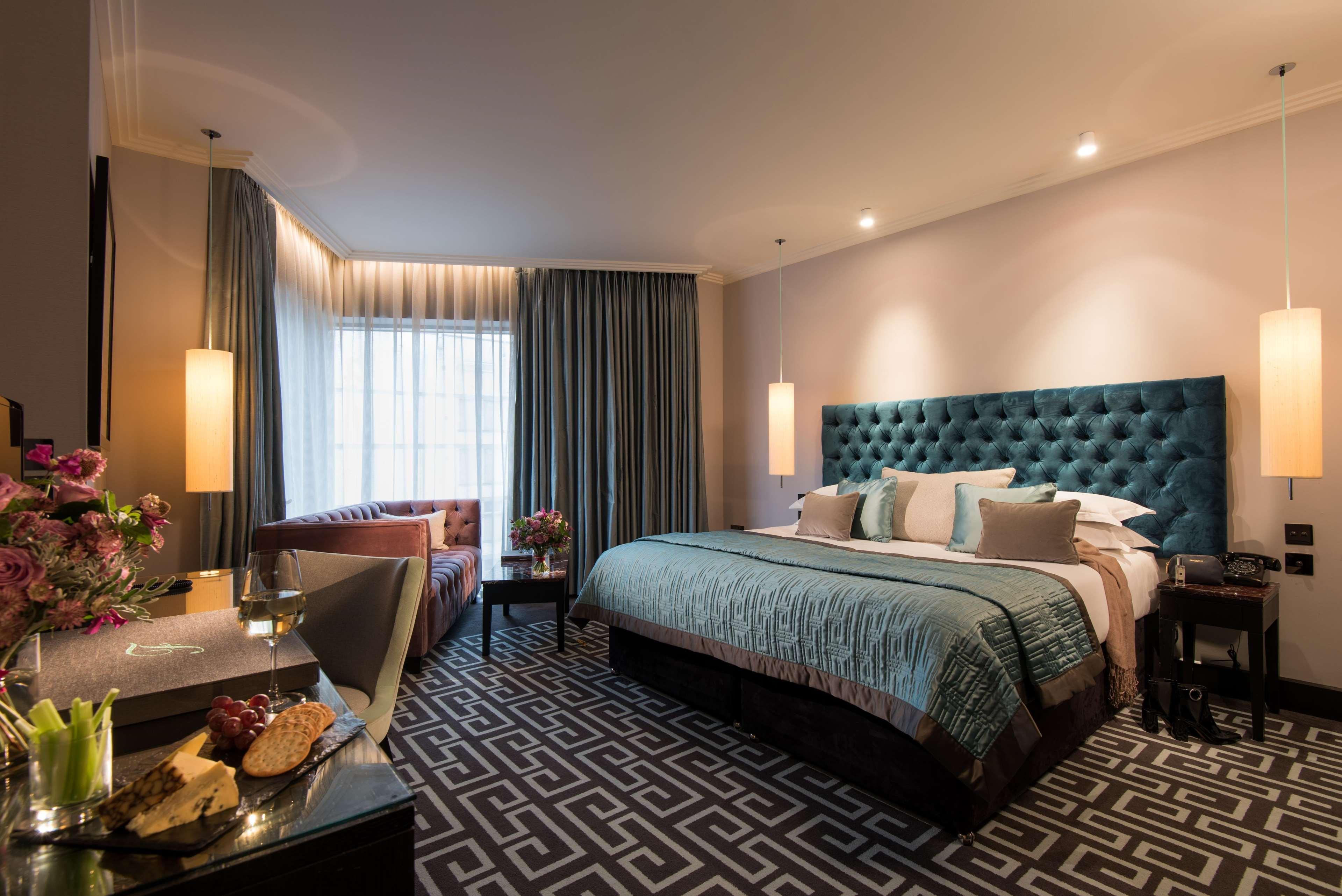 The Fitzwilliam Hotel Belfast Dış mekan fotoğraf The photo depicts a stylish hotel room. It features a large bed with a tufted teal headboard, flanked by two wall sconces providing soft lighting. The bedding is neatly arranged with accent pillows. To one side, there is a small seating area with a c