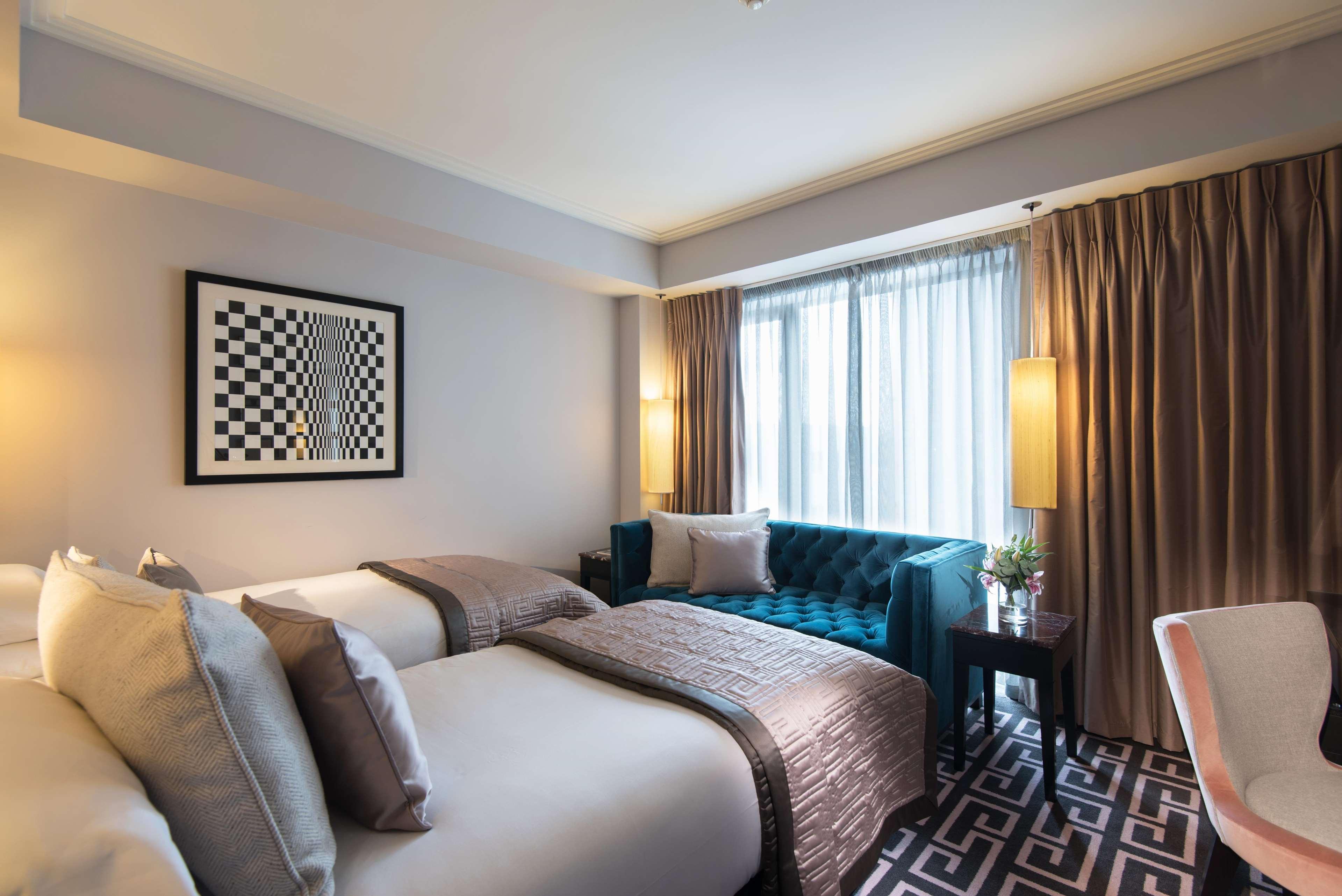 The Fitzwilliam Hotel Belfast Dış mekan fotoğraf The photo shows a modern hotel room. It features two beds with decorative pillows, a stylish teal couch, and large windows with sheer curtains allowing natural light to enter. The walls are painted in neutral tones, and there is a piece of artwork wi
