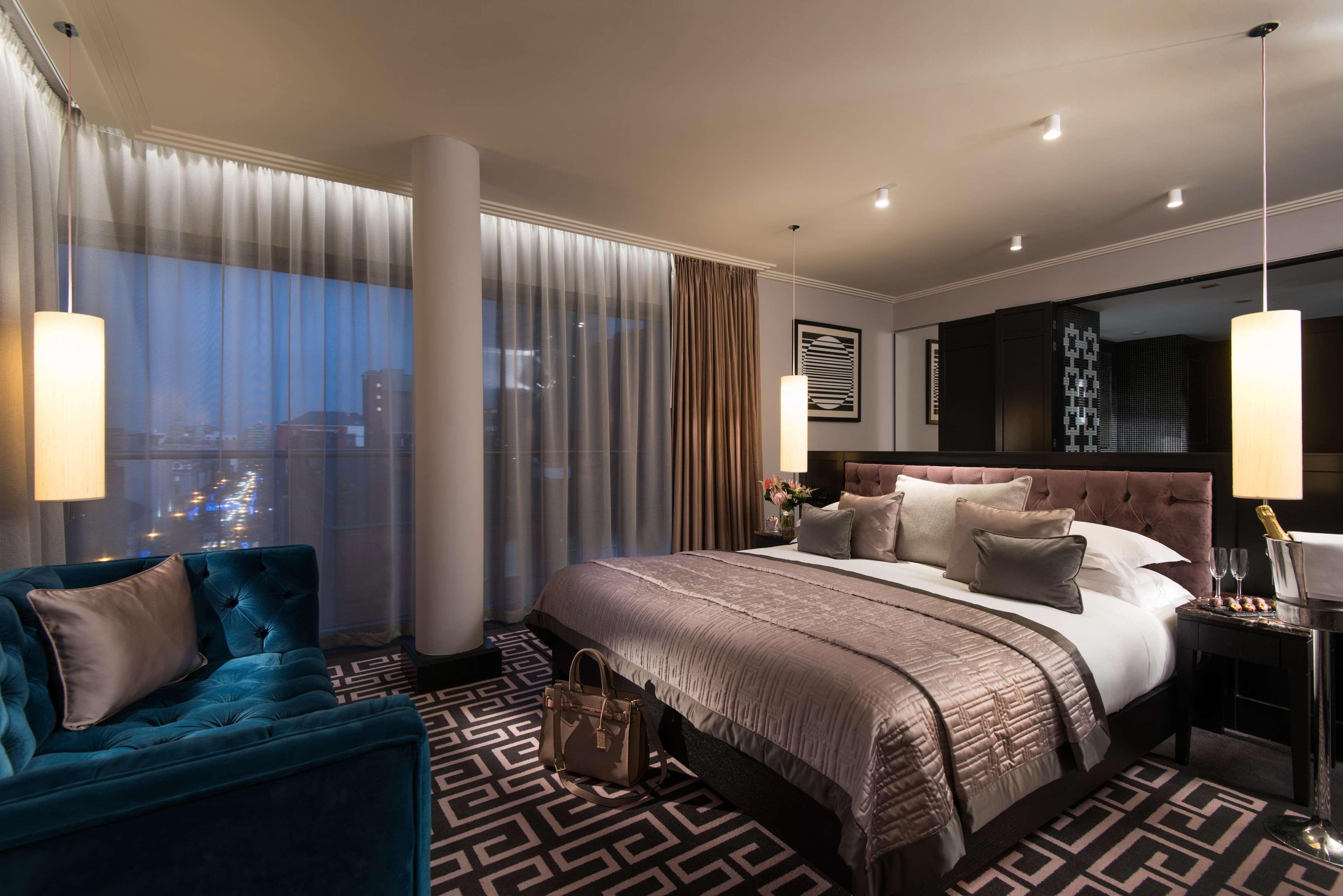 The Fitzwilliam Hotel Belfast Dış mekan fotoğraf The photo shows a luxurious hotel room with a modern design. It features a large bed with decorative pillows and a stylish bedspread, complemented by bedside lamps. To one side, there is a plush turquoise sofa and a coffee table. The room has floor-t