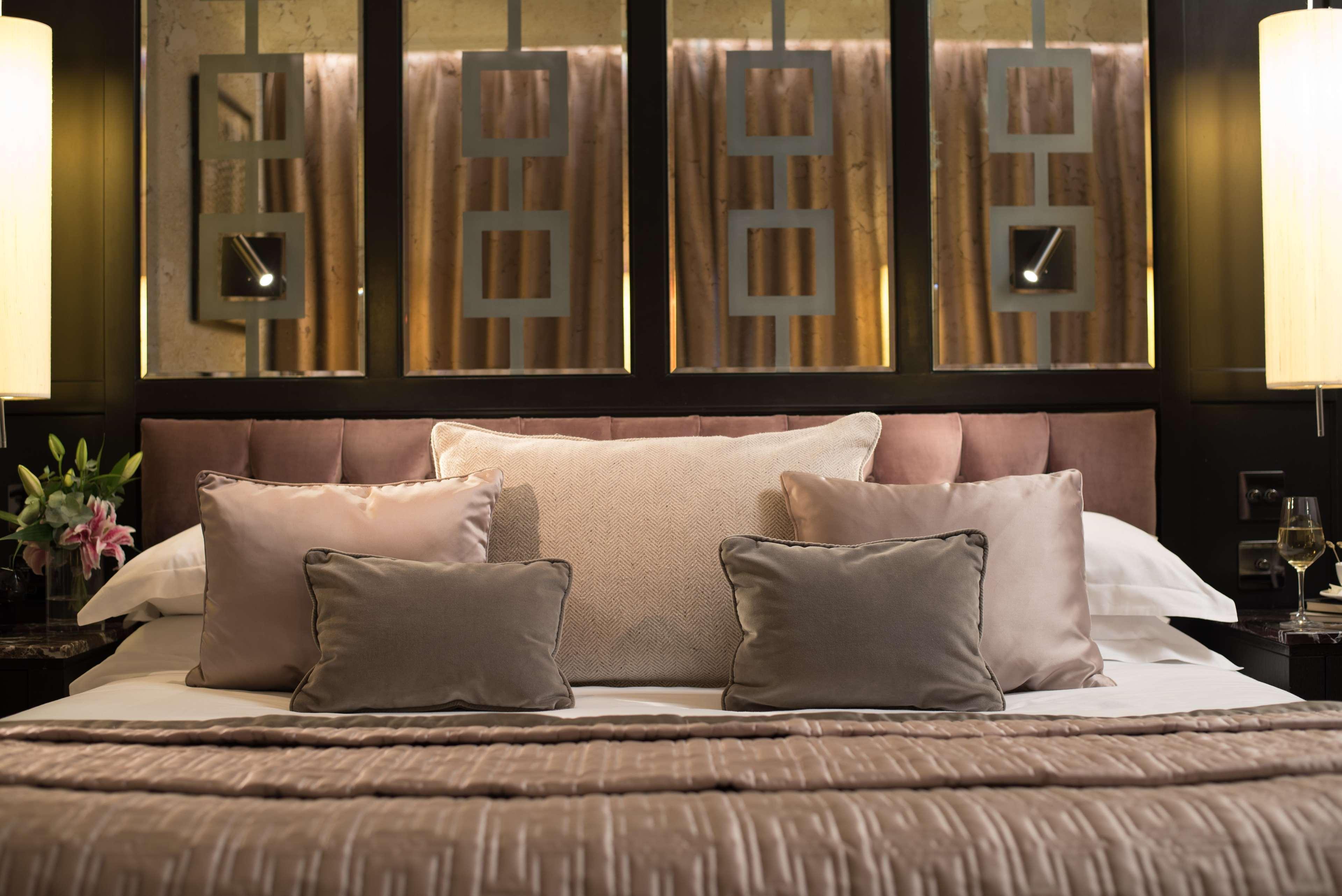 The Fitzwilliam Hotel Belfast Dış mekan fotoğraf The photo shows a beautifully designed bed in a stylish bedroom. The bed is adorned with a soft, plush headboard and layered with various decorative pillows in shades of beige and gray. The bedding appears luxurious, with a textured blanket. Behind t