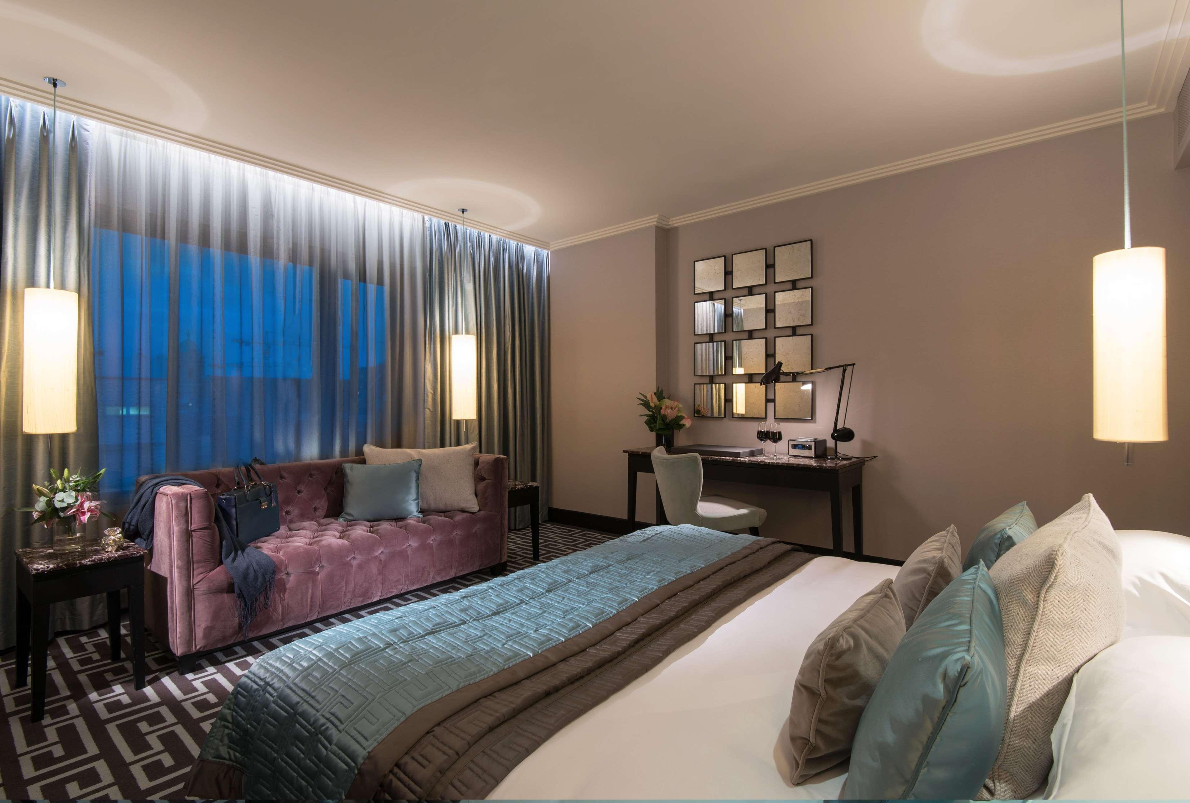 The Fitzwilliam Hotel Belfast Dış mekan fotoğraf The photo depicts a modern hotel room featuring a spacious layout. There is a large bed with decorative pillows in the foreground, covered with a stylish bedspread. To one side, a plush purple sofa adds a pop of color and comfort. A small desk and ch