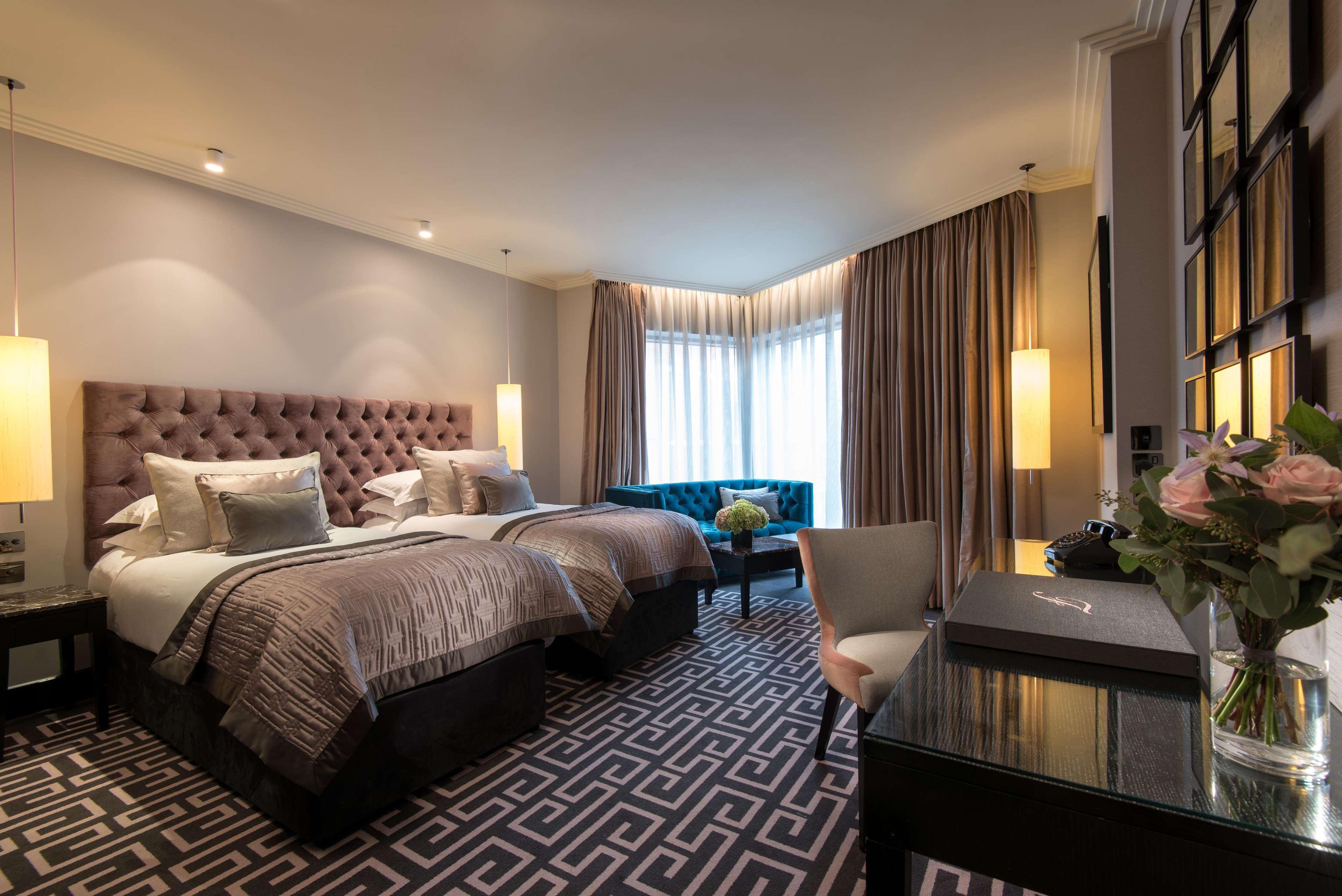 The Fitzwilliam Hotel Belfast Dış mekan fotoğraf The photo shows a stylish hotel room featuring two twin beds with tufted headboards and elegant bedding. On one side, there is a luxurious blue couch. Large windows are covered with light curtains, allowing natural light to enter. The floor is adorne