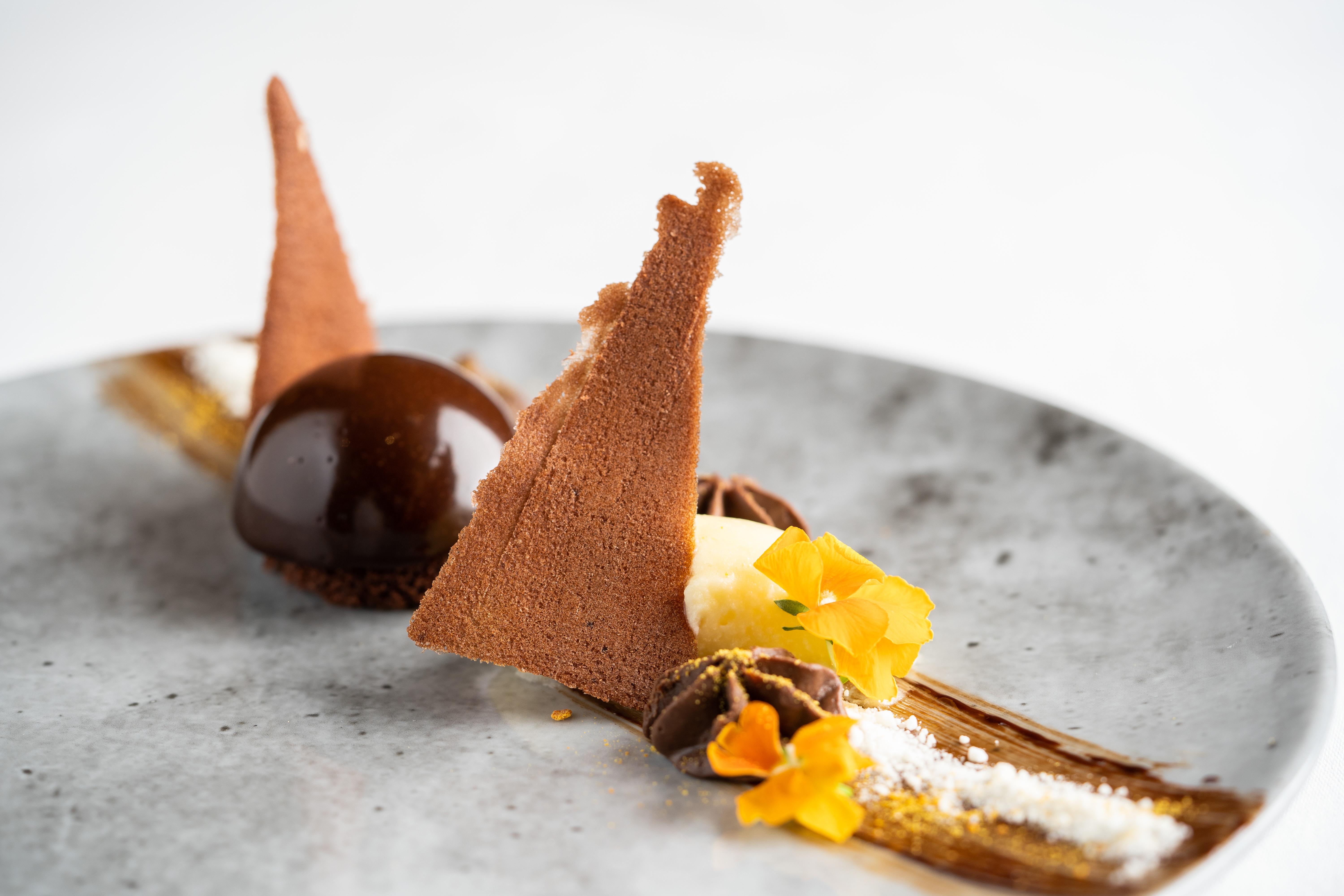 The Fitzwilliam Hotel Belfast Dış mekan fotoğraf The photo displays an artfully plated dessert. It features a glossy chocolate sphere, accompanied by various chocolate components, including triangular chocolate tuile pieces that resemble sails. The plate is adorned with delicate garnishes such as y