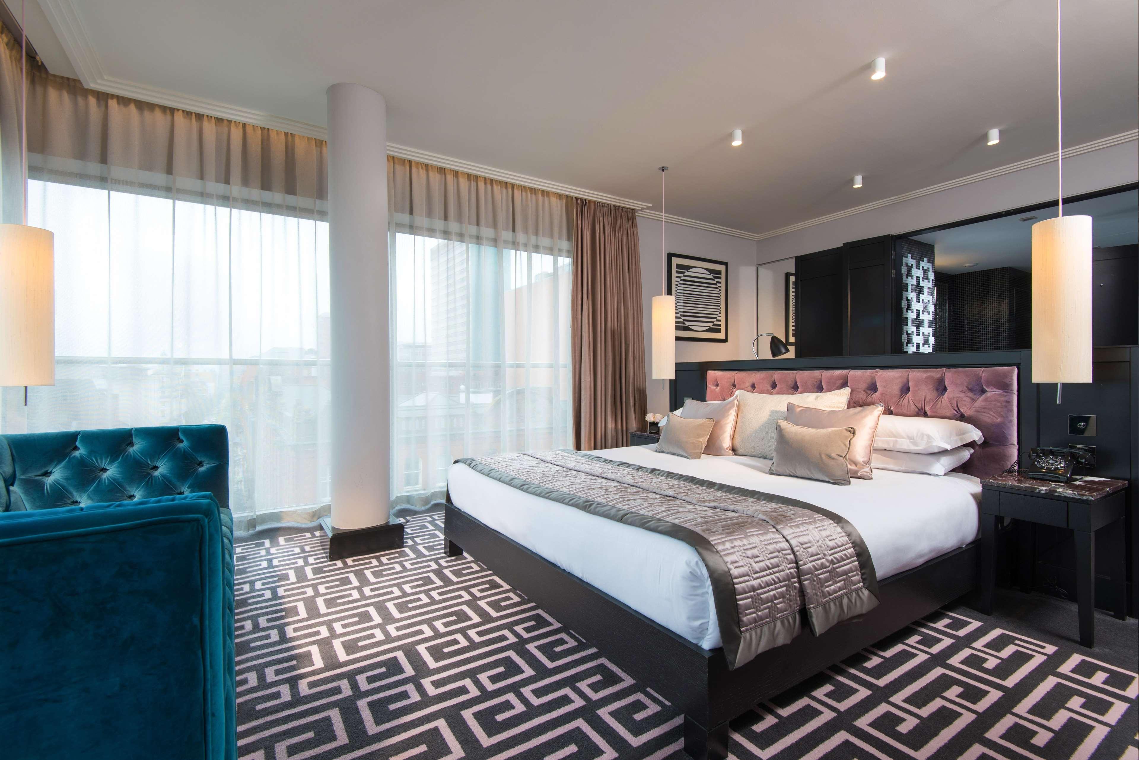 The Fitzwilliam Hotel Belfast Dış mekan fotoğraf The photo showcases a modern hotel room. It features a large bed with plush pillows and a stylish headboard. The room has large windows with sheer curtains, allowing natural light to filter in. The floor is covered with a patterned carpet that contra